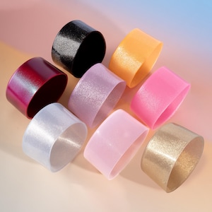 Puede incluir: Un surtido de coloridas fundas cilíndricas para bebidas en varios tonos. La colección incluye fundas negras, doradas, rosas, naranjas y moradas, cada una con un acabado ligeramente brillante. Las fundas están diseñadas para adaptarse a los recipientes de bebidas.