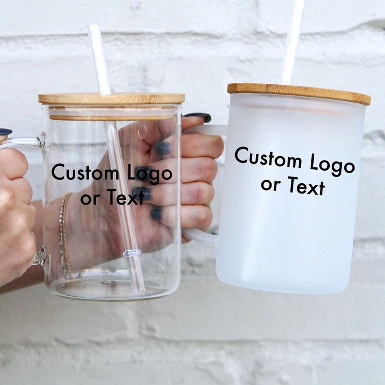 May include: Two glass mugs with bamboo lids and straws. The mug on the left is clear glass and the mug on the right is frosted glass. Both mugs have the text "Custom Logo or Text" printed on them.