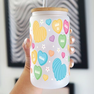 May include: White frosted glass tumbler with a bamboo lid and a straw. The tumbler has a colorful heart pattern with Spanish words like "Solo Tu" and "Dulce" on the hearts.