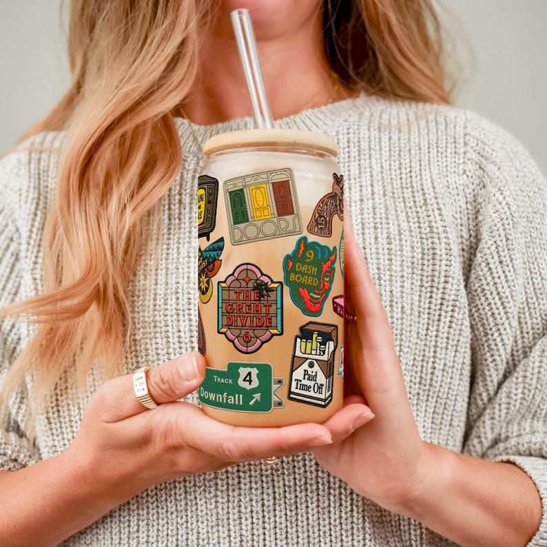 May include: A clear glass tumbler with a bamboo lid and a glass straw, decorated with vibrant stickers. The stickers display diverse designs, including text and illustrations. The tumbler is held by a person wearing a grey jumper.