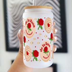 May include: White tumbler with a wooden lid and a straw. The tumbler has a design of red roses, gold stars, and pink and white pastry-like shapes. The text "de la Rosa Mazapan Original" is repeated on the tumbler.