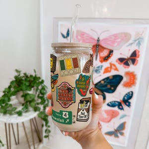 Könnte beinhalten: Ein durchsichtiger Glasbecher mit einem Holzdeckel und einem durchsichtigen Glasstrohhalm. Der Becher ist mit bunten Aufklebern bedeckt und mit einem cremigen Getränk gefüllt. Im Hintergrund befindet sich ein gerahmter Schmetterlingsdruck.