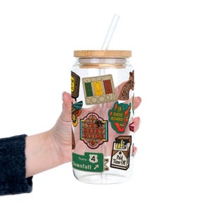 May include: A clear glass tumbler with a bamboo lid and a clear straw. The tumbler is decorated with colourful stickers, including the words "DOOR", "THE GREAT DIVIDE", "DASH BOARD", and "Paid Time Off".