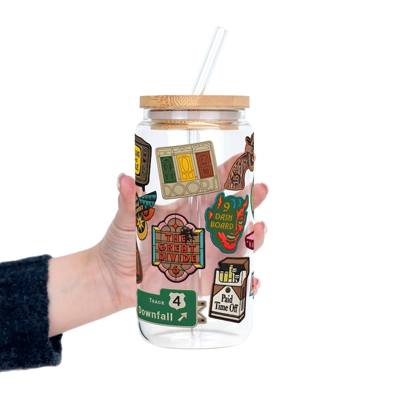 May include: A clear glass tumbler with a bamboo lid and a clear straw. The tumbler is decorated with colourful stickers, including the words "DOOR", "THE GREAT DIVIDE", "DASH BOARD", and "Paid Time Off".