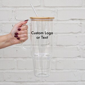 May include: A clear glass tumbler with a double wall and a bamboo lid. The tumbler has a handle and a straw. The tumbler has the text "Custom Logo or Text" printed on the side.