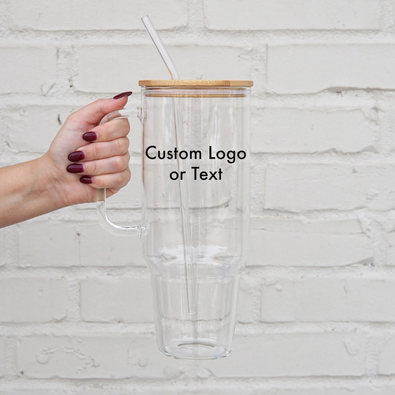 May include: A clear glass tumbler with a double wall and a bamboo lid. The tumbler has a handle and a straw. The tumbler has the text "Custom Logo or Text" printed on the side.
