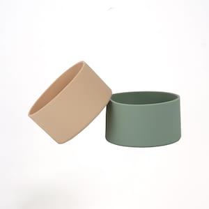 May include: Two silicone cup sleeves in neutral colors. One is a light beige, and the other is a muted green. The sleeves are cylindrical and designed to fit around a cup or bottle for insulation and protection.