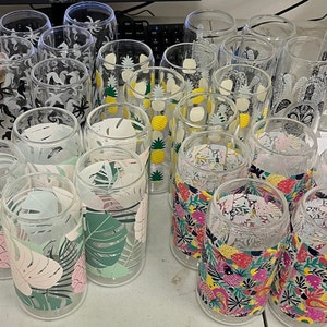 May include: A collection of clear glass tumblers with various tropical-themed designs. The designs include pineapples, palm leaves, sea turtles, and seahorses. The tumblers are arranged in a group on a white surface.