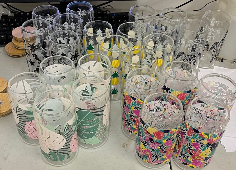 May include: A collection of clear glass tumblers with various tropical-themed designs. The designs include pineapples, palm leaves, sea turtles, and seahorses. The tumblers are arranged in a group on a white surface.
