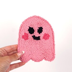 Op de afbeelding: Een roze geest met een zwart lachend gezicht en roze wangen. De geest is gemaakt van een pluche materiaal.