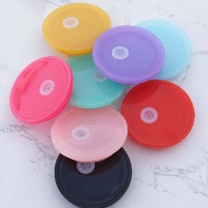 Könnte beinhalten: Eine Sammlung farbenfroher, runder Kunststoffdeckel in verschiedenen Farbtönen wie Gelb, Rosa, Lila, Blau, Rot, Grün, Schwarz und Hellrosa. Jeder Deckel hat ein zentrales, weißes, kreisförmiges Merkmal. Die Deckel sind für Tassen oder Behälter bestimmt.