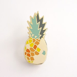Puede incluir: Un pin de piña dorado con un diseño geométrico blanco, amarillo, naranja y verde.