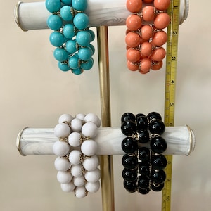 May include: Four bracelets with large, round beads. The bracelets are displayed on a marble stand. The bracelets are turquoise, coral, white, and black.