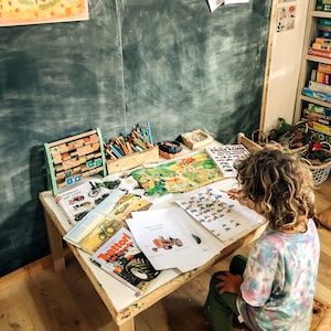 May include: A child sits at a table with a variety of educational toys and books. The table is covered in papers with pictures of tractors. The child is working on a worksheet with a picture of a tractor and the word "Tractor" written on it.