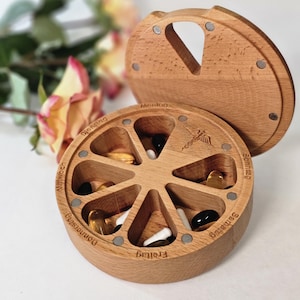 May include: A round wooden pill organizer with seven wedge-shaped compartments, each labeled with German days of the week. The lid features a cutout handle and magnetic closures. Various capsules and tablets are stored inside the compartments.