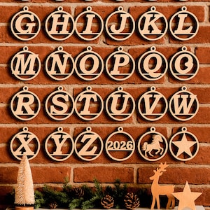 May include: Wooden alphabet ornaments, each letter suspended in a circular frame, are displayed against a brick wall. The ornaments are arranged in rows, spelling out the alphabet. A small Christmas tree, deer, and star are on a white shelf below.