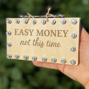 May include: A wooden money box with the text "EASY MONEY not this time" and "FUNNY GIFT MONEY BOX". The box is rectangular, with metal bolts around the edges. The background is blurred green.