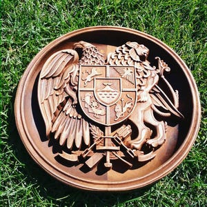 Op de afbeelding: Ronde houten plaquette met een uitgesneden wapenontwerp. De plaquette toont een adelaar, een leeuw en een centraal schild met gedetailleerde symbolen. De bruine plaquette staat tegen een achtergrond van groen gras.