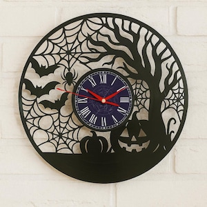 Op de afbeelding: Zwarte Halloween-thema wandklok met een cirkelvormig ontwerp. De klok heeft een spinnenweb, vleermuizen, een spin, een boom en een pompoen. De wijzerplaat is donkerblauw met Romeinse cijfers en rode wijzers.