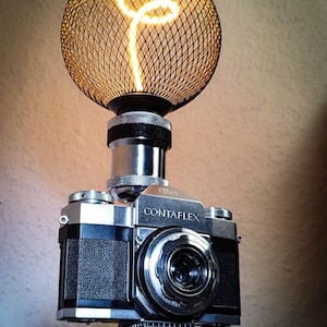 Op de afbeelding: Een unieke lamp met een vintage Contaflex camerabody, bekroond met een bolvormige draadkooi met een gloeiende gloeilamp. De lamp staat op een miniatuur Eiffeltoren basis, met het woord "PIRANIUM" op een klein plaatje.