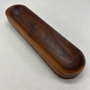 May include: A long, oval-shaped wooden dish made of dark walnut wood. The dish has a smooth, rounded interior and a slightly raised edge.