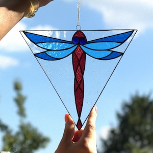 Peut inclure: Un vitrail en forme de libellule dans un cadre triangulaire. La libellule a des ailes bleues et un corps rouge, avec des contours noirs. La pièce est suspendue par une fine ficelle, sur fond de ciel bleu et de feuillage vert.