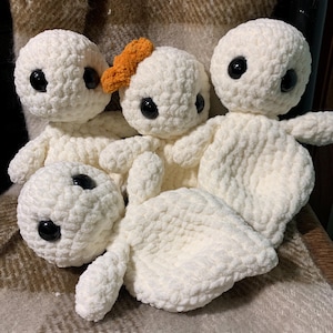 May include: A collection of five handmade, white crochet dolls with black button eyes. One doll has an orange crochet accessory. The dolls are arranged on a textured, brown and cream-colored blanket.