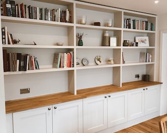 Può includere: Librerie a muro bianche con un piano di lavoro in legno e armadietti bianchi sotto. Gli scaffali sono pieni di libri e oggetti decorativi.