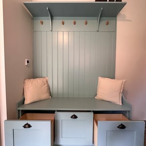 May include: A light blue wooden bench with three drawers and a shelf above. The bench has two beige cushions and black drawer pulls. The shelf has wooden hooks and is holding a pair of brown shoes.