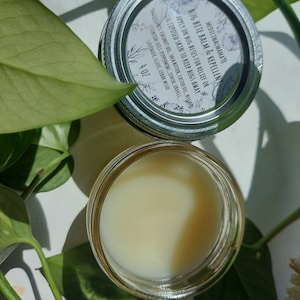 May include: A small glass jar with a white label that reads "Medicinal Mamas Bug Bite Balm & Repellent". The label also includes instructions for use and a list of ingredients. The jar is filled with a light yellow balm.