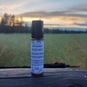 May include: A roll-on bottle labeled "Medicinal Mama Bug Bite Roller" with a black cap. The label includes text about relieving bug bites and ingredients. The bottle is 10 ml and sits on a wooden surface with a scenic background.