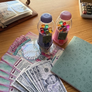May include: Two gumball machine banks, one purple and one pink, filled with colorful gumballs. A stack of US five-dollar bills and a budget planner are also visible. The image also includes a Gumball Savings Machine game.