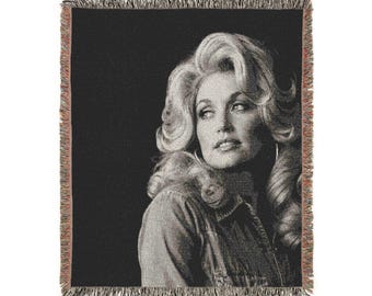 May include: A woven throw blanket featuring a black and white portrait of a person with voluminous blonde hair. The blanket has a dark background, a textured fabric construction, and a decorative fringed edge along all four sides of the rectangular piece.