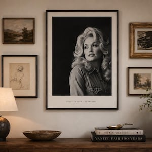 May include: A large framed black and white portrait of Dolly Parton centered on a wall, surrounded by four smaller framed artworks. The portrait includes the text Dolly Parton - Tennessee at the bottom. A lamp, bowl, and books sit on the table below.