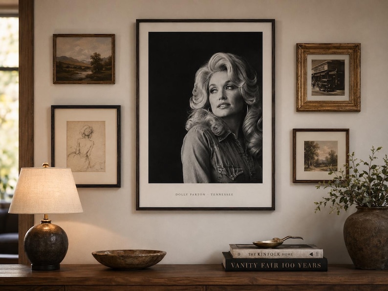 May include: A large framed black and white portrait of Dolly Parton centered on a wall, surrounded by four smaller framed artworks. The portrait includes the text Dolly Parton - Tennessee at the bottom. A lamp, bowl, and books sit on the table below.