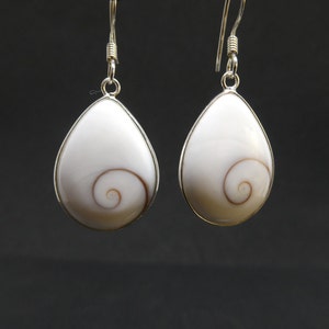 Op de afbeelding: Een paar druppelvormige oorbellen met witte Shiva-oorschelpen met een natuurlijk bruin spiraalpatroon. Elke schelp is gezet in een zilverkleurige metalen rand en hangt aan een eenvoudige zilverkleurige oorhaak voor doorboorde oren.