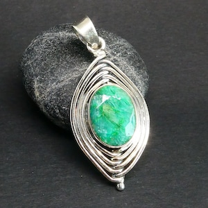 May include: A silver pendant featuring an oval-shaped, faceted green gemstone set in the centre. The metal frame consists of multiple concentric, wire-like silver loops that form an elongated, pointed oval shape around the central stone.
