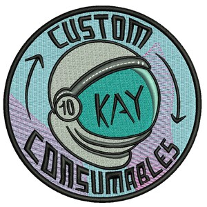 May include: A round, blue and pink embroidered patch with a white astronaut helmet and the text "CUSTOM 10 KAY CONSUMABLES" in black.