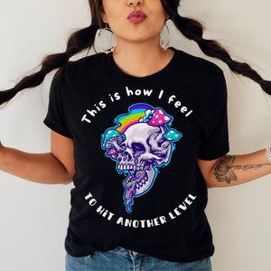 May include: Black t-shirt with a white skull graphic and rainbow mushrooms. The text "This is how I feel to hit another level" is printed on the shirt.