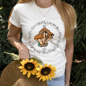 May include: White t-shirt with a graphic of brown mushrooms in a circle of black vines. The shirt has a short sleeve and a crew neck.