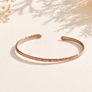 May include: A hammered copper cuff bracelet. The open-ended bracelet has a textured surface and a warm, reddish-brown color. The bracelet is displayed on a neutral background with soft shadows.