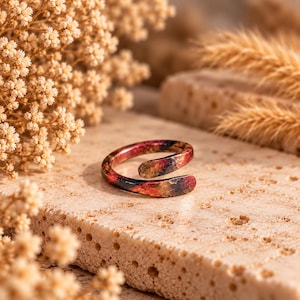 May include: A bypass ring featuring a hammered texture with a gradient color finish transitioning from deep black to vibrant red and orange tones. The metal band has an open design with overlapping ends, showcasing a metallic sheen throughout.