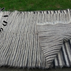 Puede incluir: Una manta de lana a rayas con un esquema de color blanco y gris. La manta está doblada por la mitad y tiene un borde con flecos.