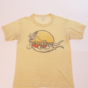 May include: A faded yellow t-shirt featuring the word "imagine" in a stylized font, with a graphic of a bird and a sun. The design is in shades of orange, red, and black. The shirt has short sleeves and a crew neck.