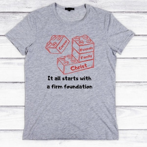 以下が含まれることがあります： 積み木を積み重ねた赤いグラフィックが描かれたグレーのTシャツ。積み木には「キリスト」、「家族」、「友人」、「キャリア」とラベルが付けられています。「すべては堅固な基盤から始まる」というテキストが積み木の下に印刷されています。