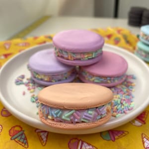 May include: A plate of colourful macarons with pastel-coloured shells and rainbow sprinkles. The macarons are in shades of pink, purple, and peach. The plate sits on a yellow cloth with ice cream cone designs.