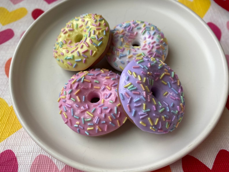 Pode incluir: Quatro donuts coloridos com cobertura pastel e granulado arco-&iacute;ris est&atilde;o dispostos em um prato branco. Os donuts s&atilde;o amarelos, rosa, branco e roxo. O fundo &eacute; uma toalha de mesa com estampa de cora&ccedil;&atilde;o.