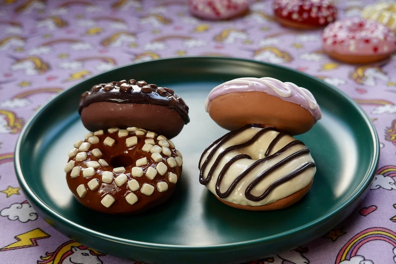 Pode incluir: Quatro donuts decorados em um prato verde. Dois est&atilde;o empilhados, um com cobertura de chocolate e granulado branco, o outro com cobertura de chocolate. Os outros dois donuts t&ecirc;m cobertura branca e chocolate.