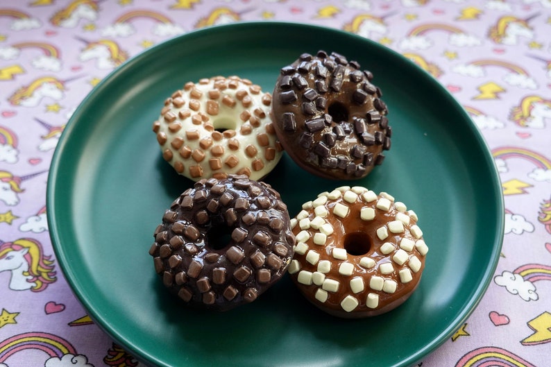 Pode incluir: Quatro donuts de chocolate com v&aacute;rias coberturas, incluindo gotas de chocolate e quadrados de chocolate branco, est&atilde;o dispostos em um prato verde escuro. Os donuts est&atilde;o em uma mesa com um padr&atilde;o de unic&oacute;rnios e arco-&iacute;ris.