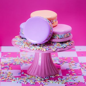May include: A stack of three round, smooth macarons in pastel purple, peach, and pink, filled with white cream and colorful sprinkles, resting on a pink pedestal cake stand covered in scattered multi-colored sprinkles against a pink background.